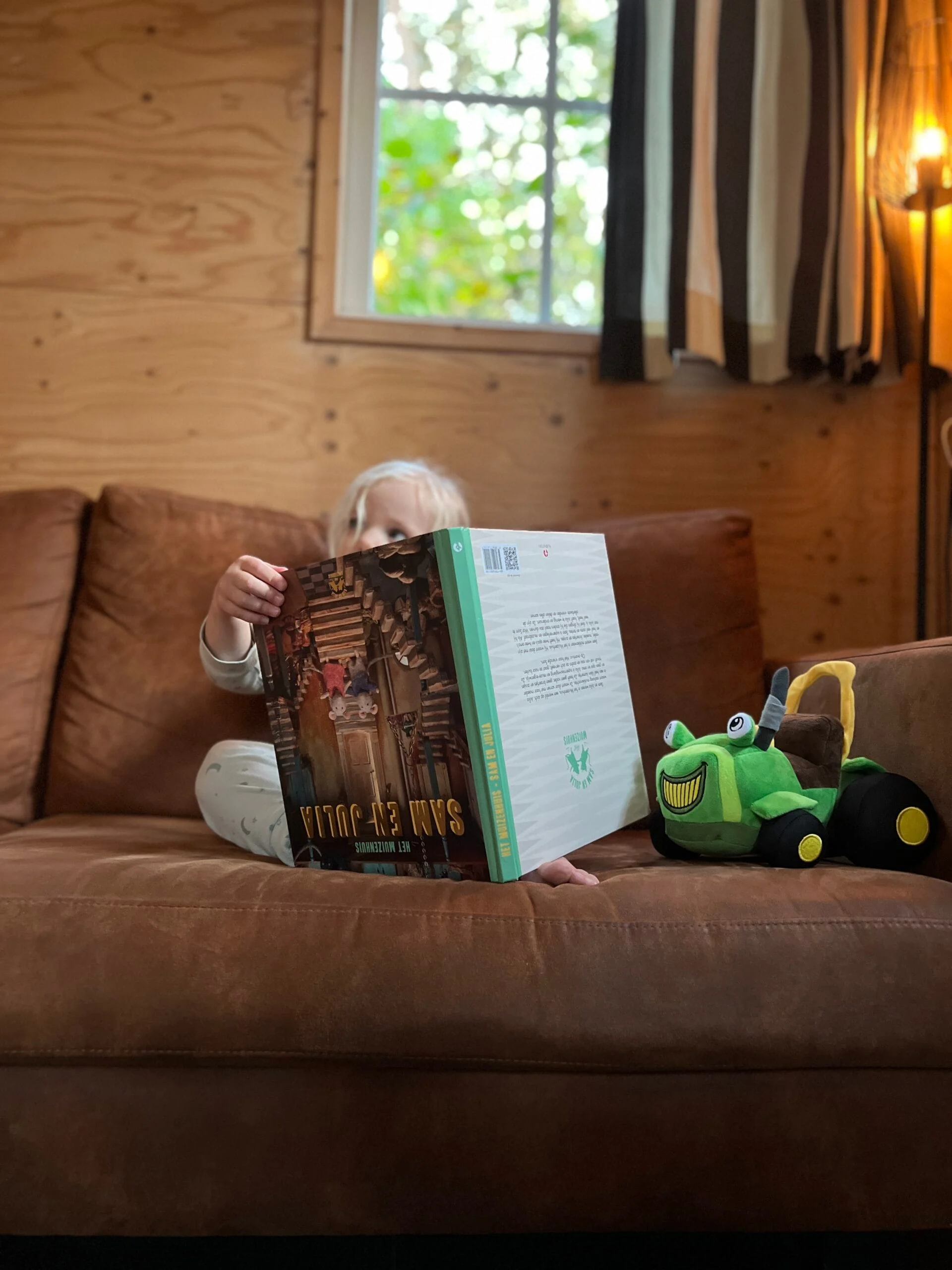 Kind leest een boek op de bank in De Hooikiep met een tractor knuffel van de Norgerberg ernaast