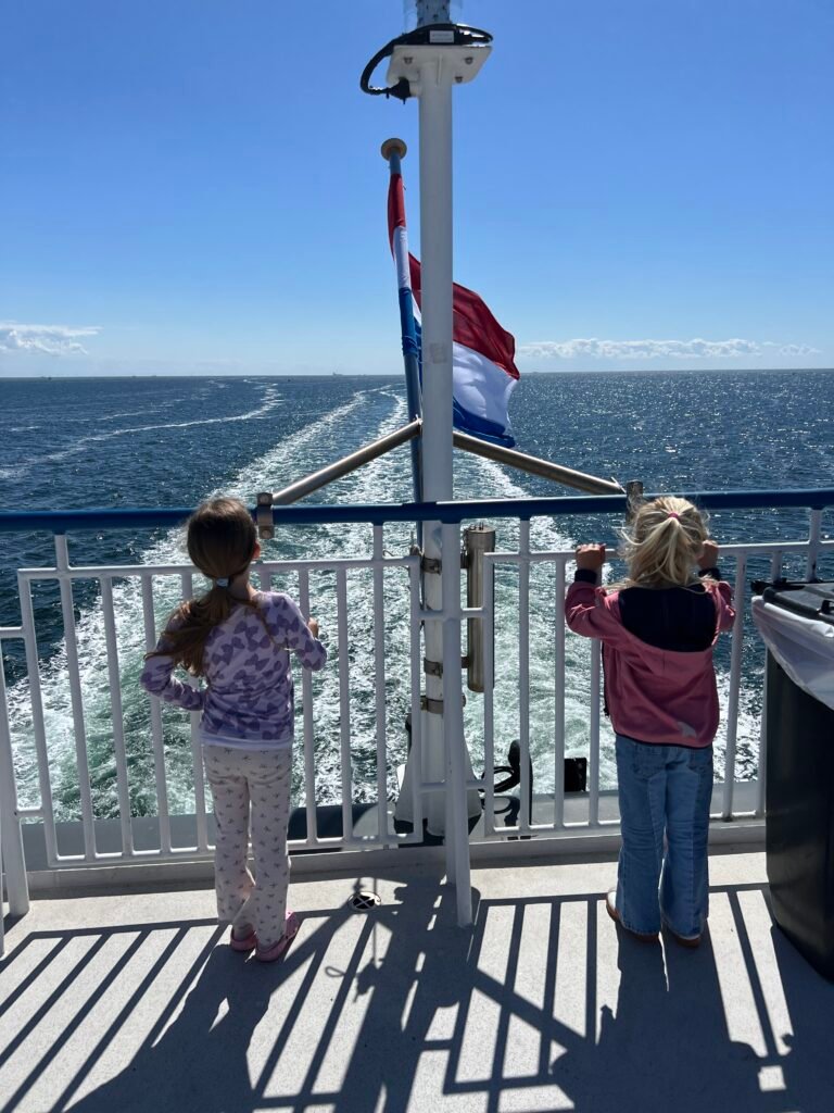 Kinderen kijken vanaf het achterdek van de veerboot uit over de Waddenzee met de Nederlandse vlag in beeld