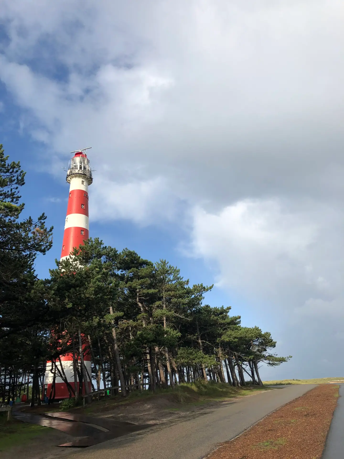 Rood-wit gestreepte vuurtoren van Ameland steekt boven de bomen uit onder een blauwe lucht met wolken